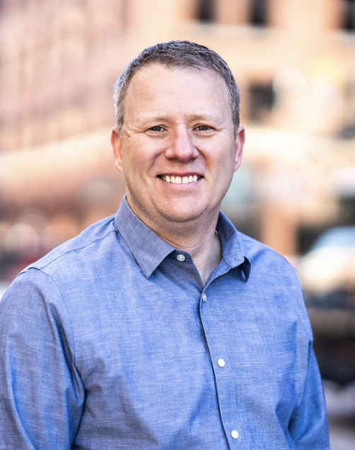 A smiling man with short light brown hair wearing a blue button-up shirt, standing outdoors in an urban setting with blurred buildings and cars in the background.