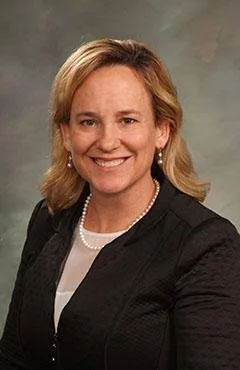 A professional woman with blonde hair, wearing a black blazer and pearl necklace, smiling for a portrait.