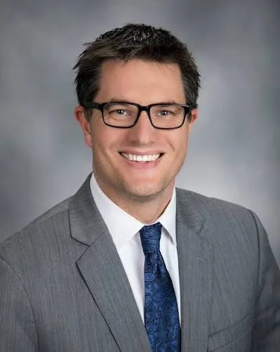 A smiling man with dark hair, glasses, wearing a gray suit, white shirt, and blue tie, posing against a gray background.