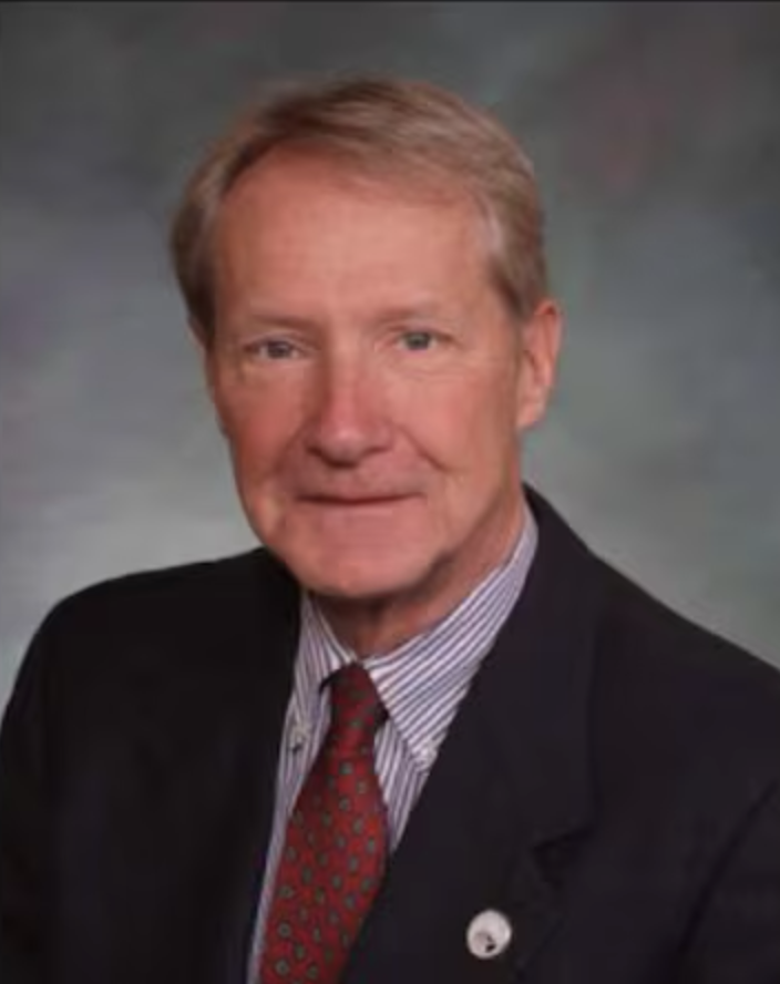 A professional portrait of an older man with blonde hair wearing a dark suit, a striped shirt, and a red patterned tie, against a gray background.