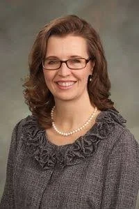 A woman with shoulder-length brown hair, glasses, and a pearl necklace, wearing a gray sweater with ruffle detailing at the collar, smiling against a neutral background.