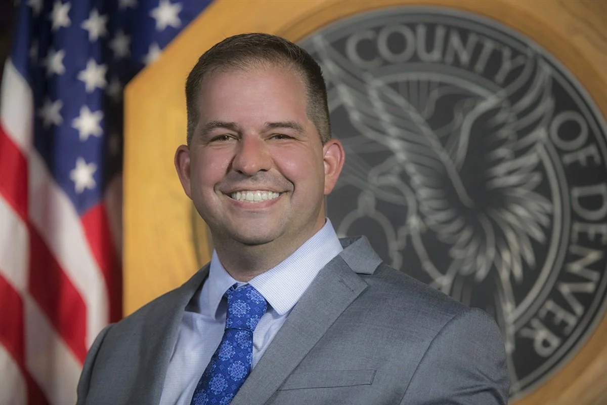 A man in a gray suit with a light blue shirt and blue patterned tie, smiling, standing in front of a US flag and a government seal.