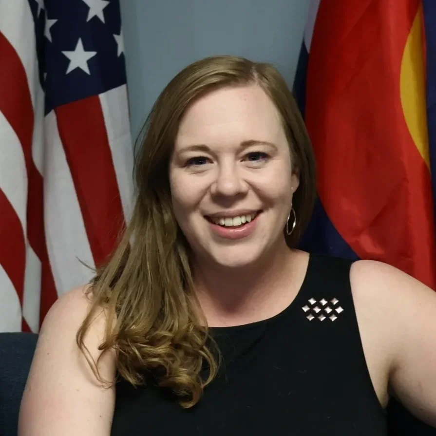A woman with long red hair smiling in front of American and other flags.
