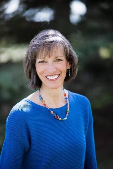 A smiling woman with short brown hair and earrings, wearing a blue sweater and a colorful necklace, outdoors with blurred natural background.