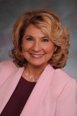 A smiling woman with shoulder-length, styled blonde hair, wearing a light pink blazer over a dark top, and a silver necklace, against a neutral background.