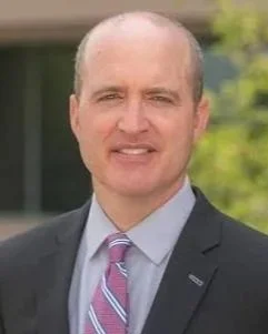 A man with short hair and a receding hairline, dressed in a suit and tie, standing outdoors with a blurred background of greenery and a building.