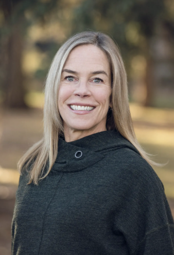 A woman with blonde hair smiling outdoors in a park with trees in the background.