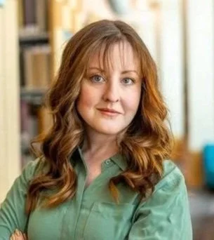 A woman with wavy, shoulder-length hair and a green shirt standing indoors near bookshelves.