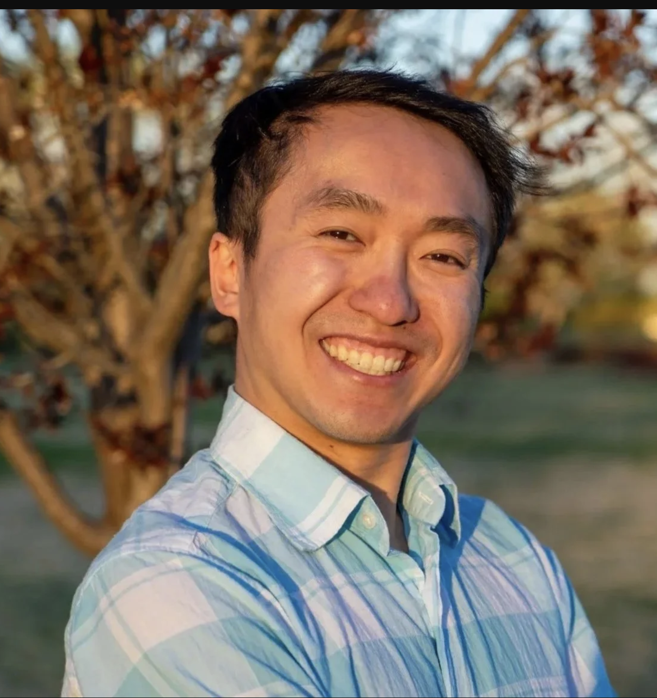A smiling man in a blue and white striped shirt standing outdoors with a tree in the background during sunset.