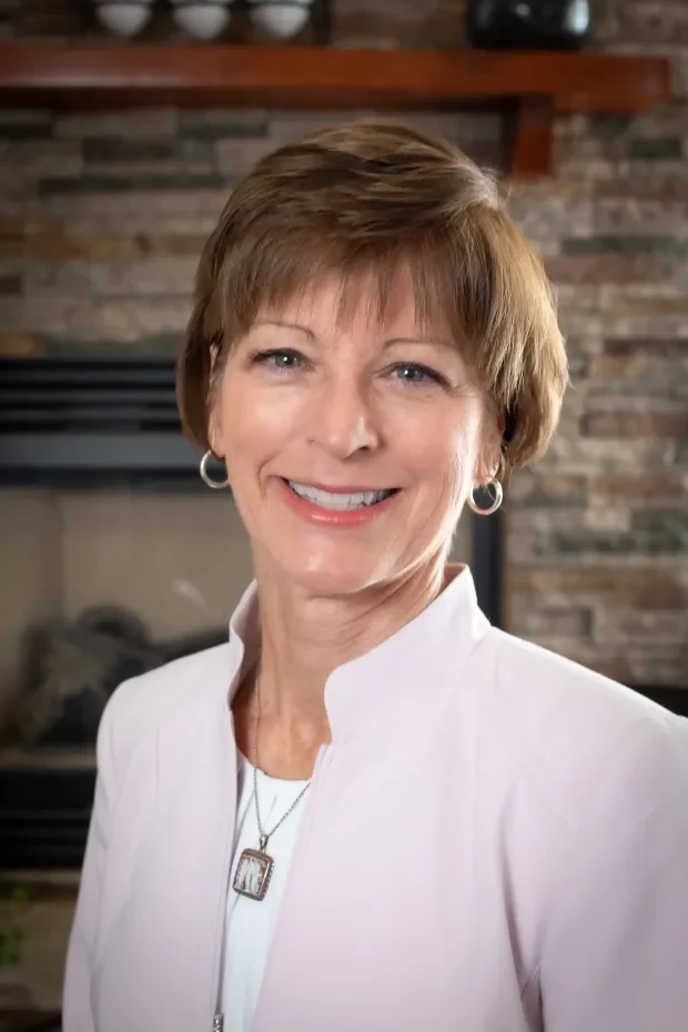 A smiling middle-aged woman with short brown hair, wearing a white blazer, earrings, and a necklace, standing indoors in front of a brick fireplace.