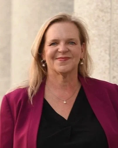 Portrait of a woman with blonde hair, wearing a black top and a maroon blazer, standing outdoors with a neutral background.