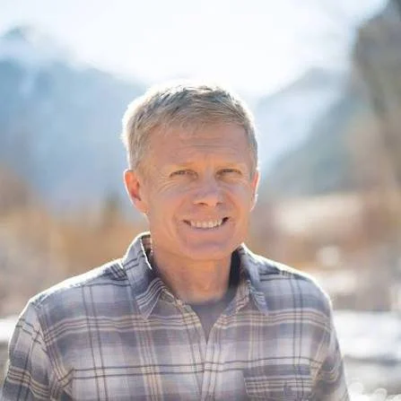 A smiling middle-aged man with short gray hair, wearing a plaid shirt, outdoors in a mountain area with snow.