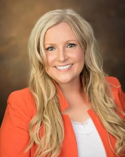 A smiling woman with long blonde hair wearing an orange blazer over a white top, posing against a brown background.