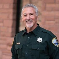 A smiling man dressed as a sheriff with a badge, insignia, and a dark uniform, standing outdoors against a brick wall background.