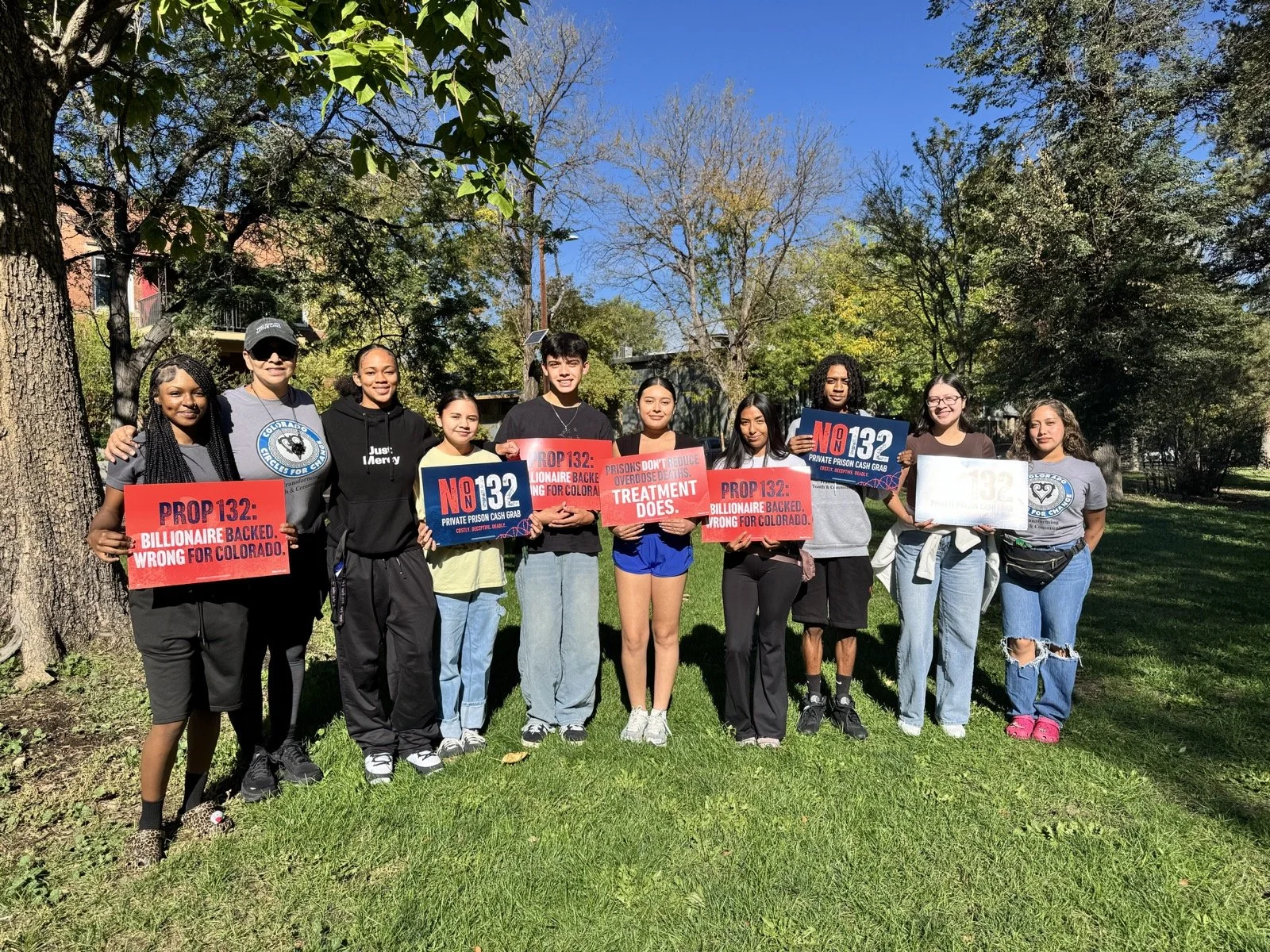Group of diverse young people standing outdoors on grass, holding protest signs against Prop 132, with trees and a blue sky in the background.