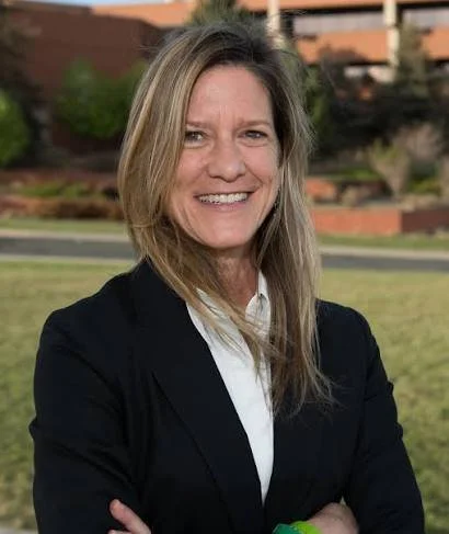 A woman with long blonde hair smiling outdoors in front of a landscaped background wearing a black blazer and white shirt.