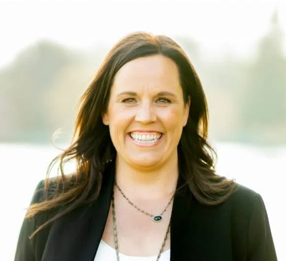 A woman with long dark hair smiling outdoors, wearing a black blazer and a necklace.