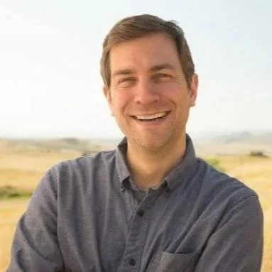 A man smiling outdoors in a field with a blue sky in the background.