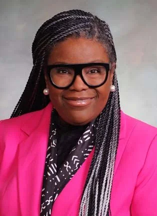 A woman with braided hair, wearing glasses, pearl earrings, a pink blazer, and a black and white patterned scarf against a plain background.