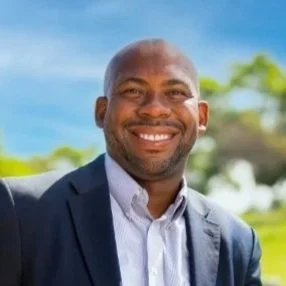 A smiling man in a suit outdoors on a sunny day with trees and blue sky in the background.