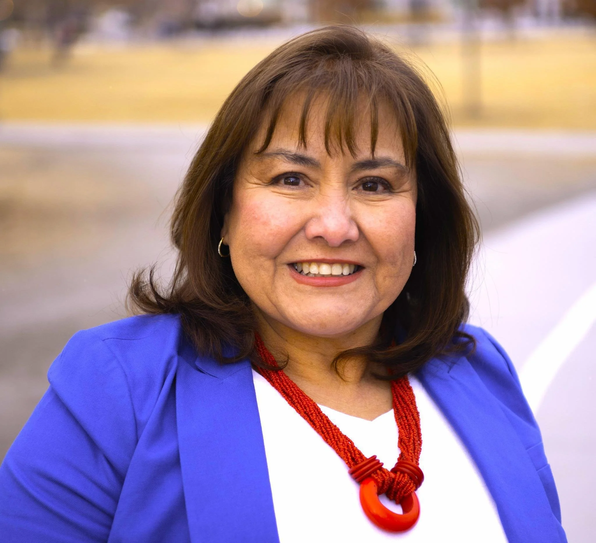 A woman with brown hair, wearing a blue blazer, white shirt, and red beaded necklace, smiling outdoors.