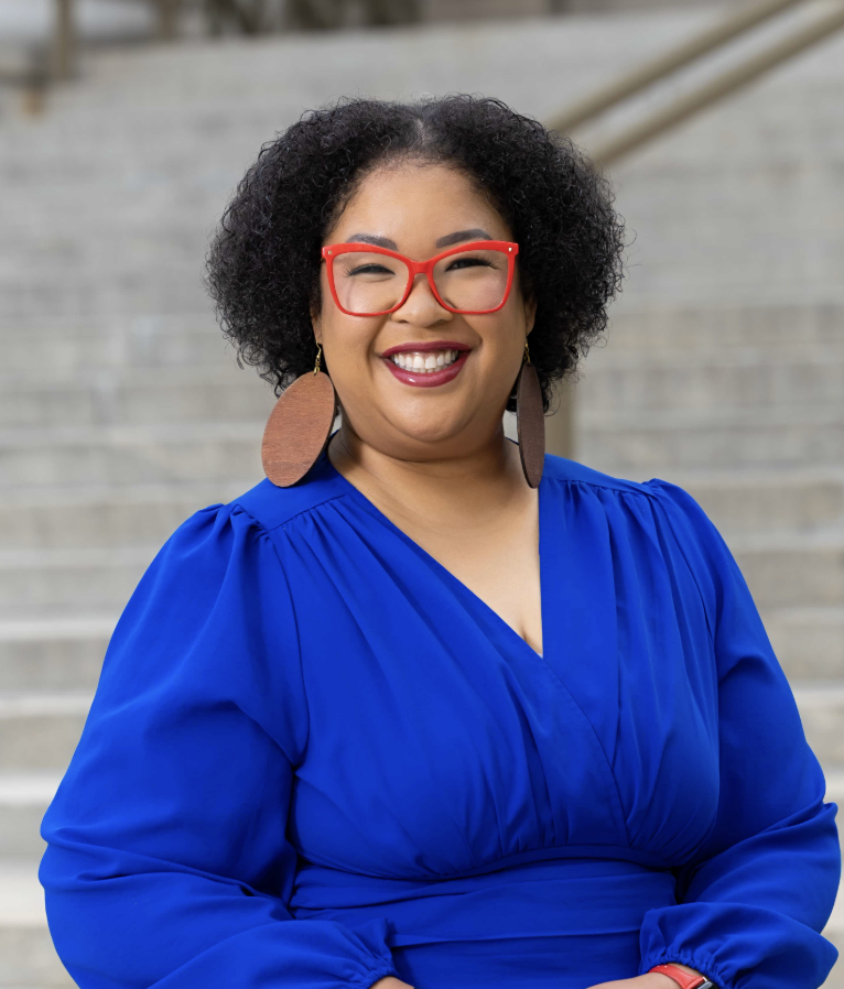 A smiling woman wearing red glasses, large wooden earrings, a blue dress, and standing outdoors on stairs.
