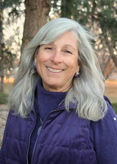 A smiling woman with long gray hair wearing a purple jacket outdoors, with trees in the background.