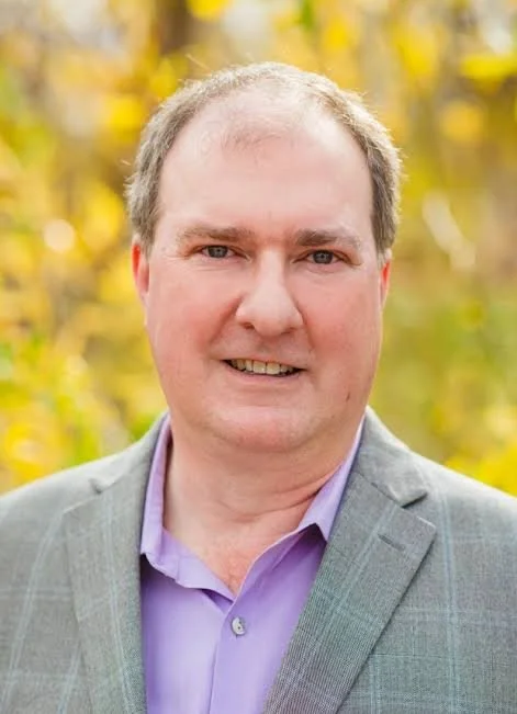 Portrait of a man in a gray blazer and lavender shirt smiling outdoors with yellow autumn leaves in the background.