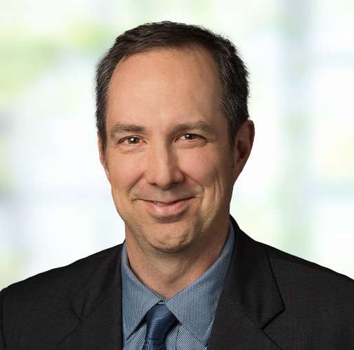 Headshot of a man in a suit and tie smiling at the camera, with a blurred office background.
