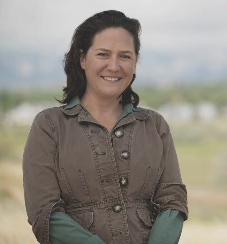 A woman smiling outdoors, wearing a brown jacket with rolled-up sleeves, and a green shirt underneath. Blurred landscape background with sky and distant mountains.