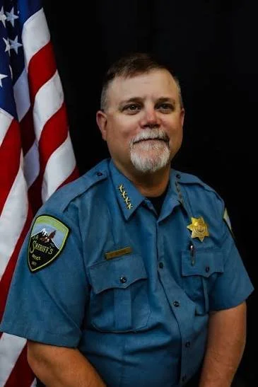 A male police officer standing in front of a United States flag, wearing a blue uniform with a sheriff's badge and patches.