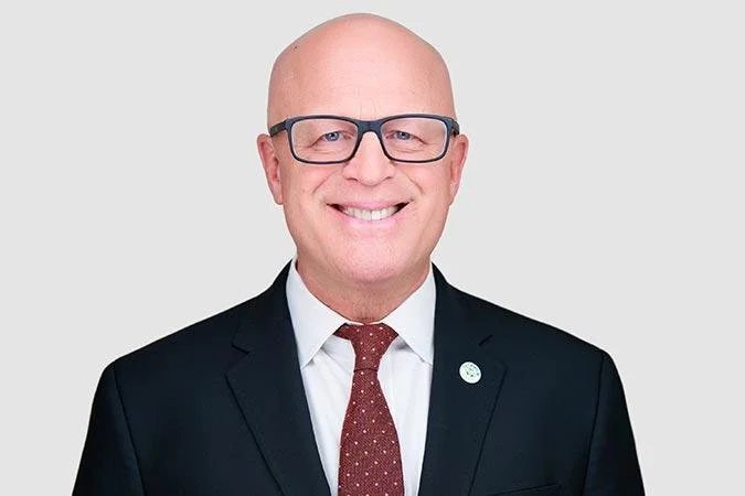 A man in a black suit, white shirt, and red patterned tie, smiling at the camera against a plain white background.