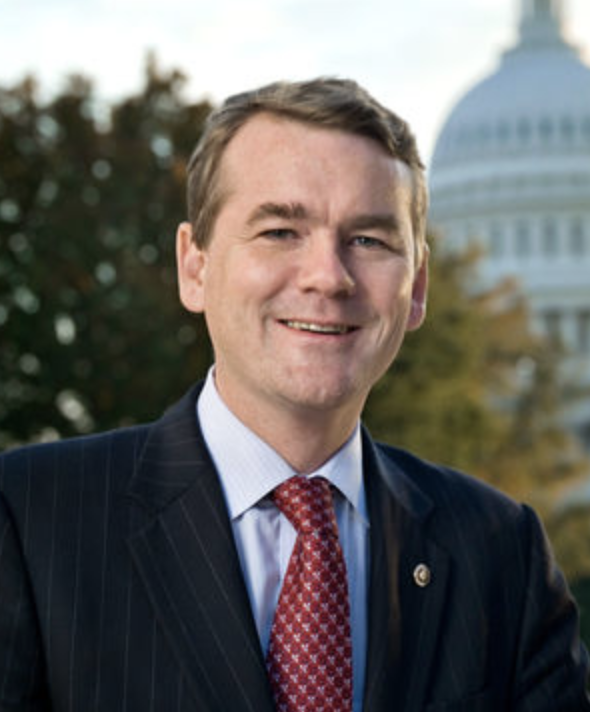 A smiling man in a business suit, standing outdoors with the Capitol building in the background.