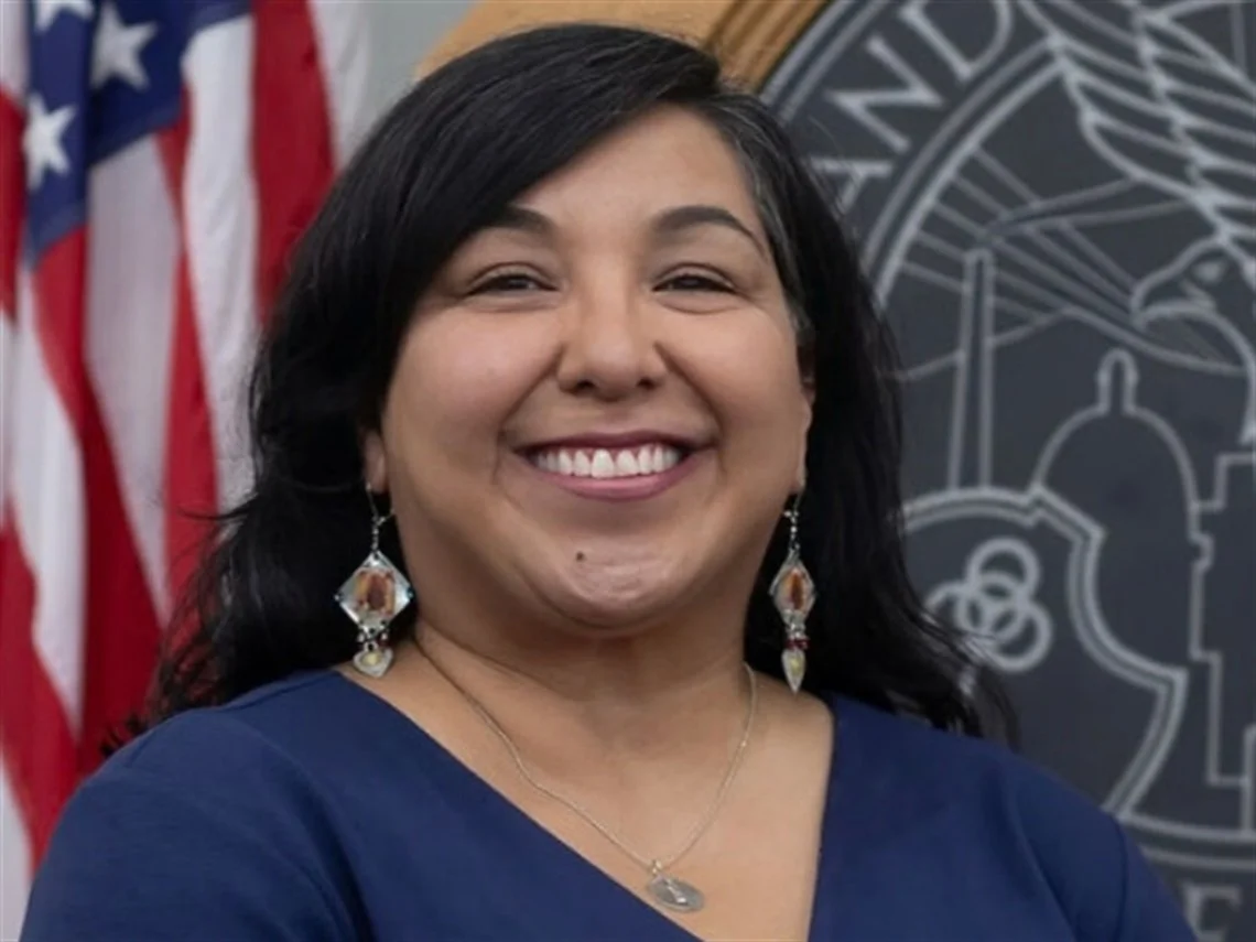A woman smiling, standing in front of American flags and an emblem of a government or official building.