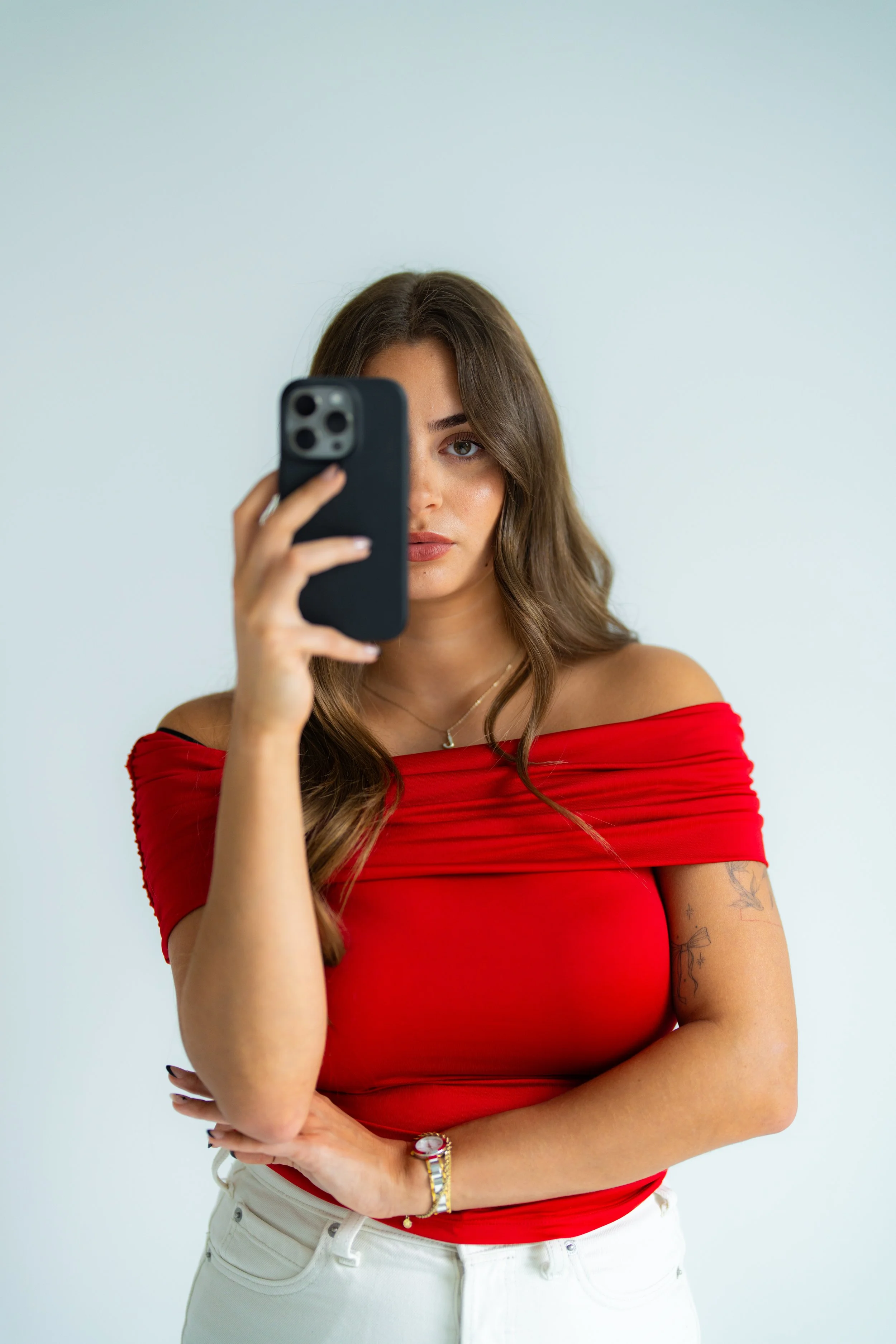A young woman with long brown hair and tattoos on her right arm, wearing an off-the-shoulder red top and white pants, taking a selfie with a smartphone.