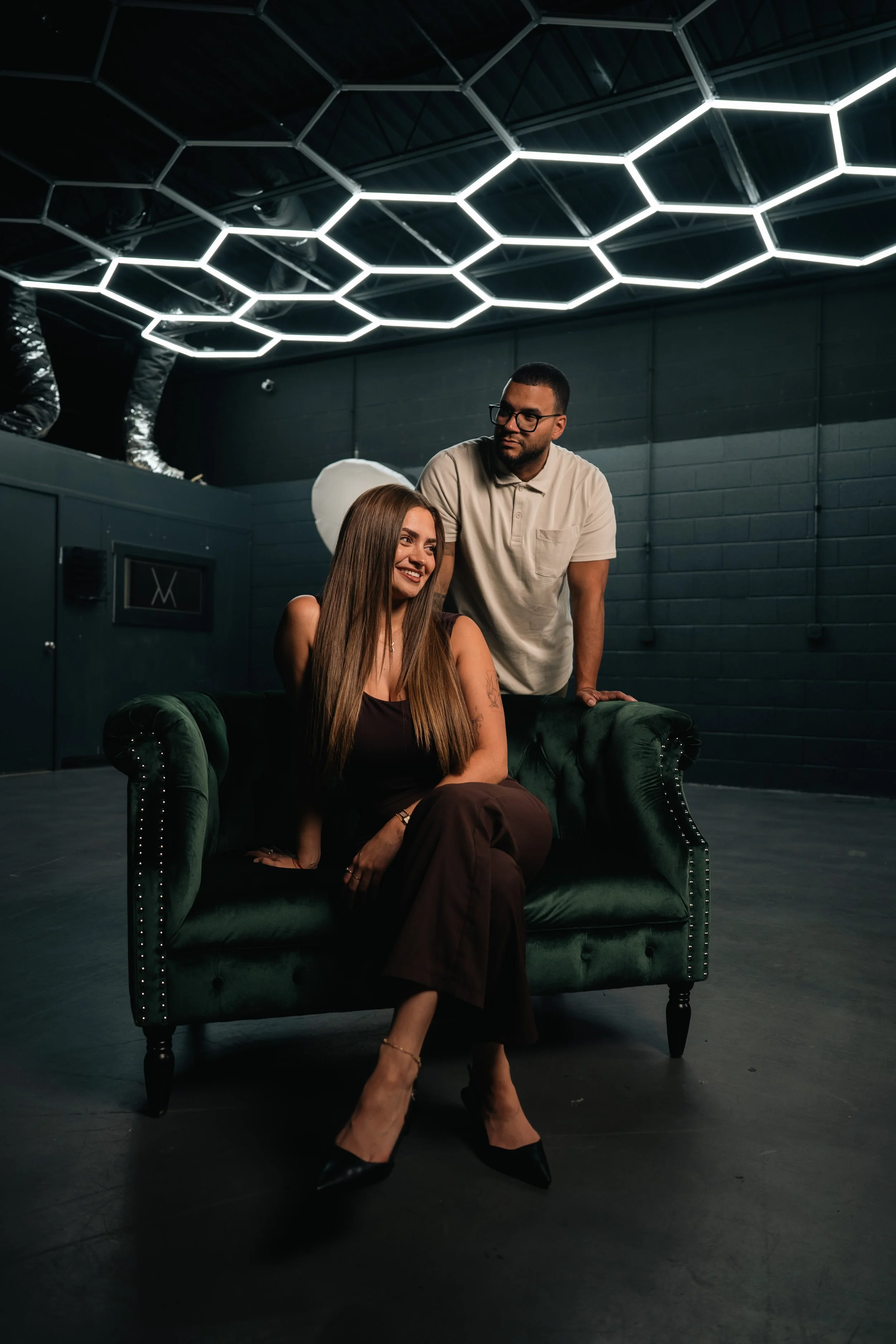A woman with long brown hair sits on a dark green velvet tufted sofa with high heels. A man with glasses and short dark hair stands behind her, resting his hand on the back of the sofa. They are in a room with dark walls and a ceiling with a modern honeycomb-shaped light fixture.
