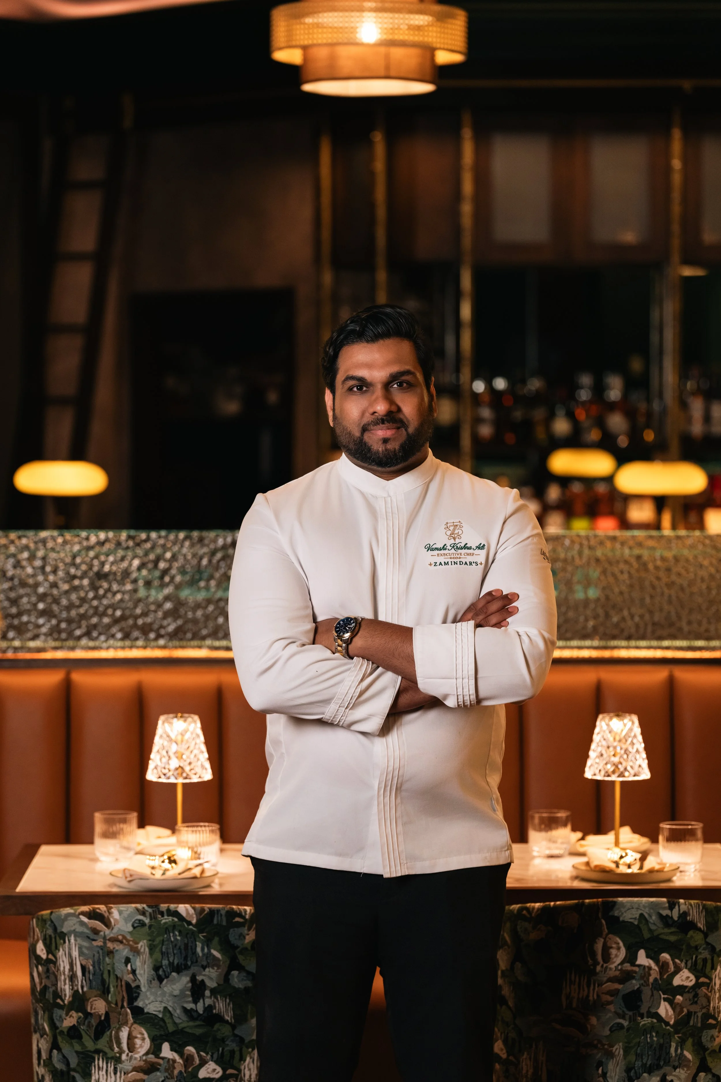 A man dressed in a white chef's coat standing with arms crossed in a restaurant setting, with a bar and dining tables with lamps in the background.