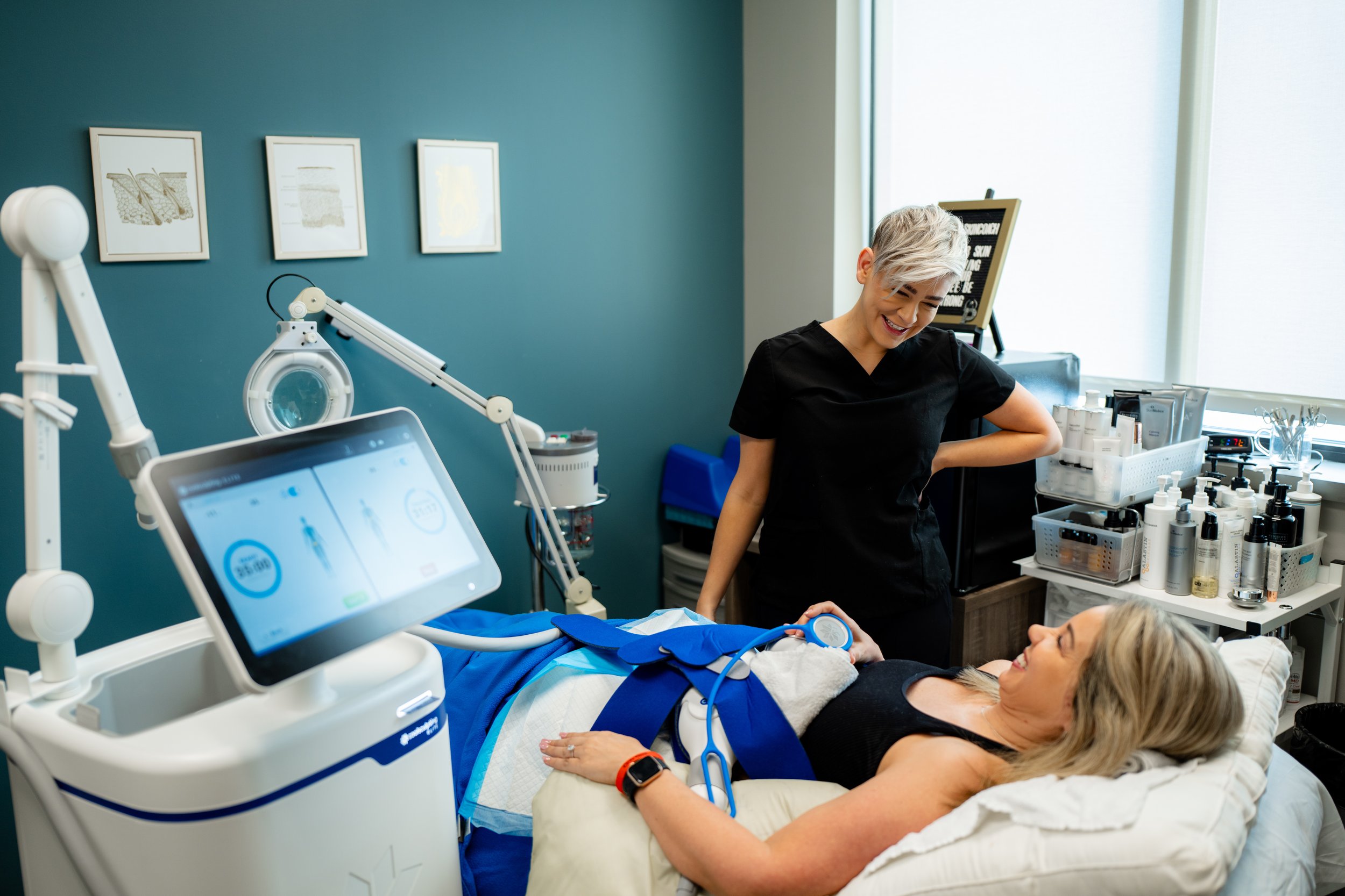 A woman lying in a hospital bed with medical equipment around her, talking to a healthcare professional and smiling, in a clinical setting with a teal wall and framed artwork.