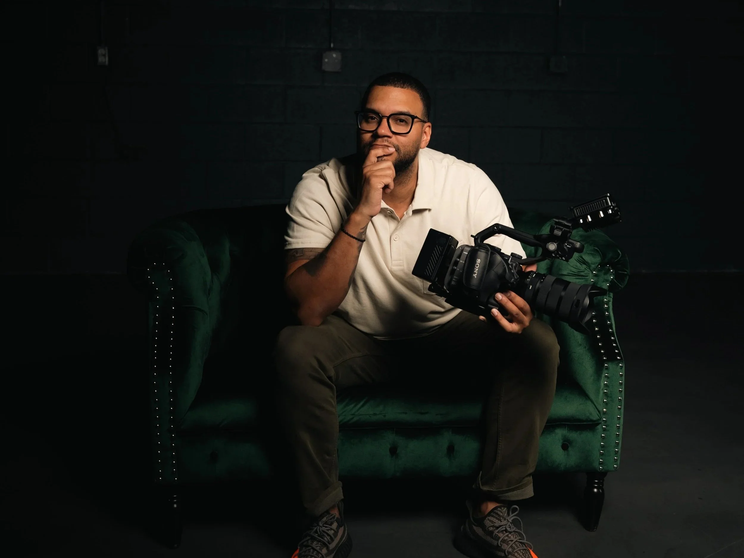 A man with glasses and a beard sits on a green velvet couch in a dark room, holding a professional video camera, with his hand on his chin.