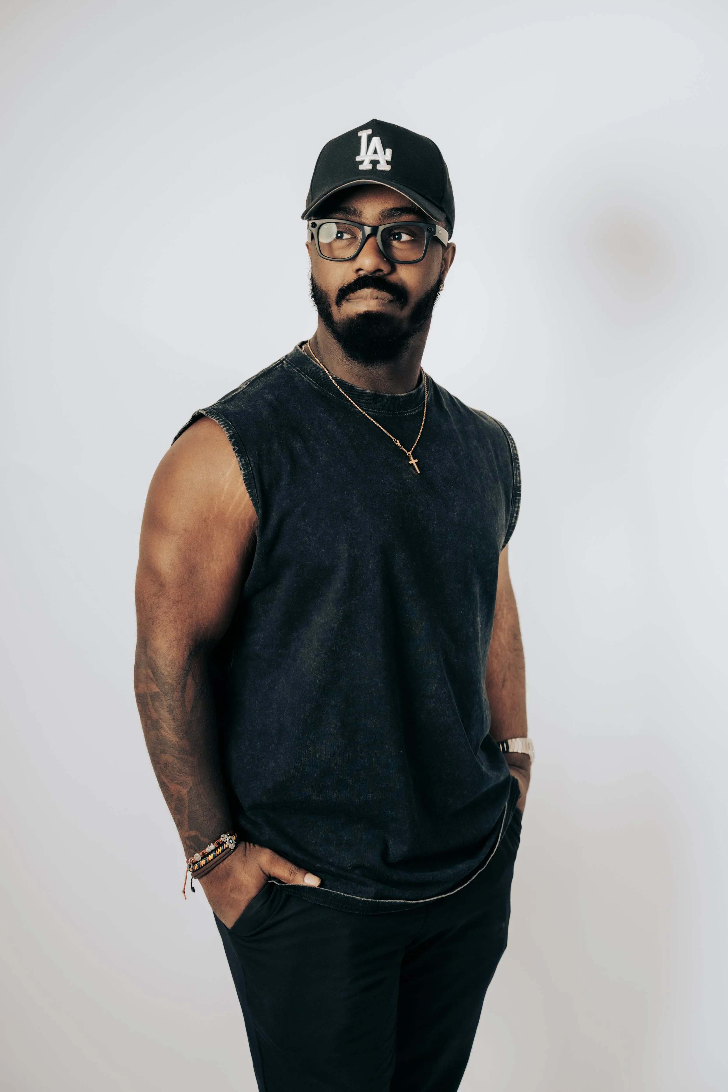 A man wearing a black LA baseball cap, glasses, a sleeveless black shirt, and black pants, standing with one hand in his pocket against a plain white background, accessorized with necklaces and bracelets.