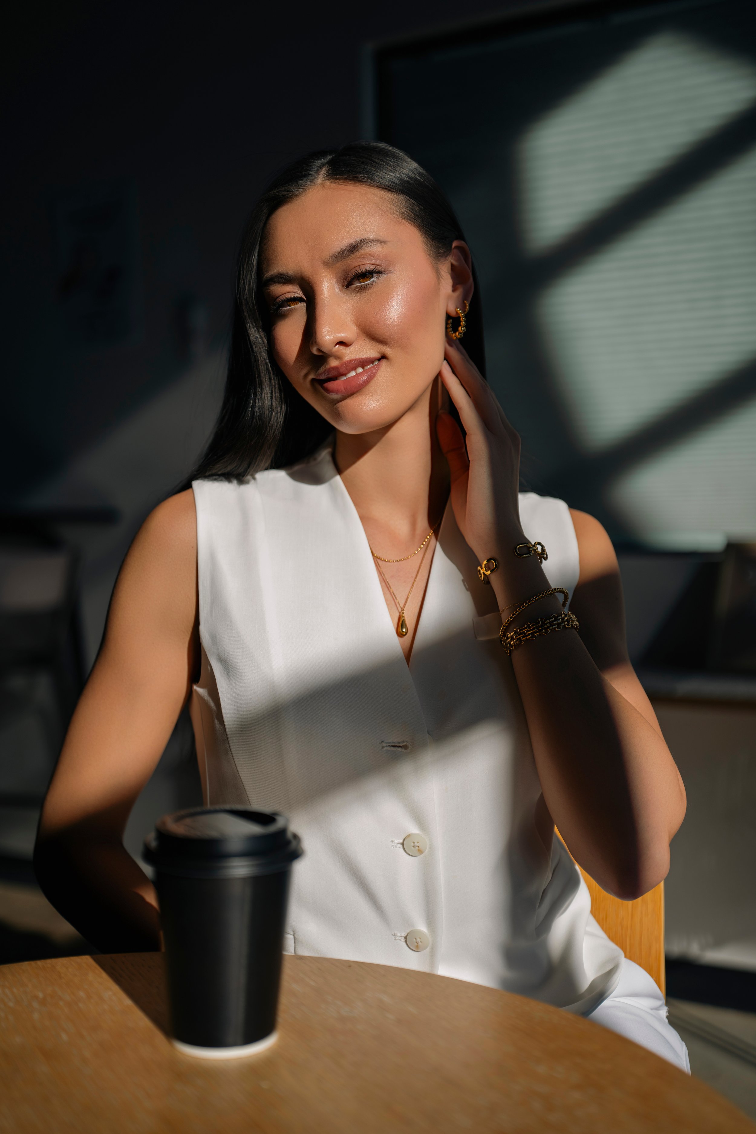 A woman with dark hair and gold jewelry sitting at a wooden table, touching her neck, with sunlight creating shadows through blinds in the background, and a coffee cup in the foreground.