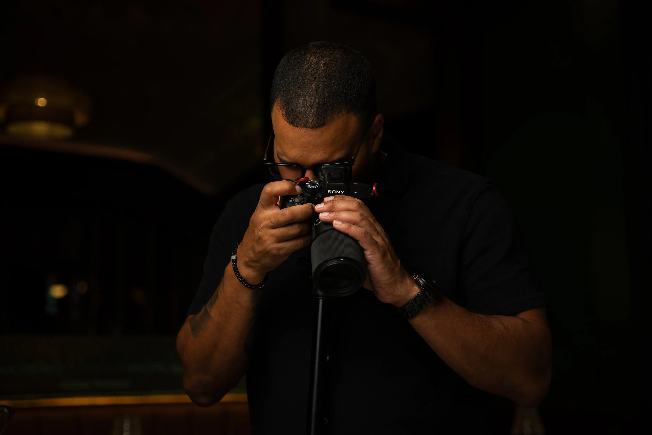 A person with short dark hair and glasses holding a Sony camera, looking through the viewfinder in a dark environment.