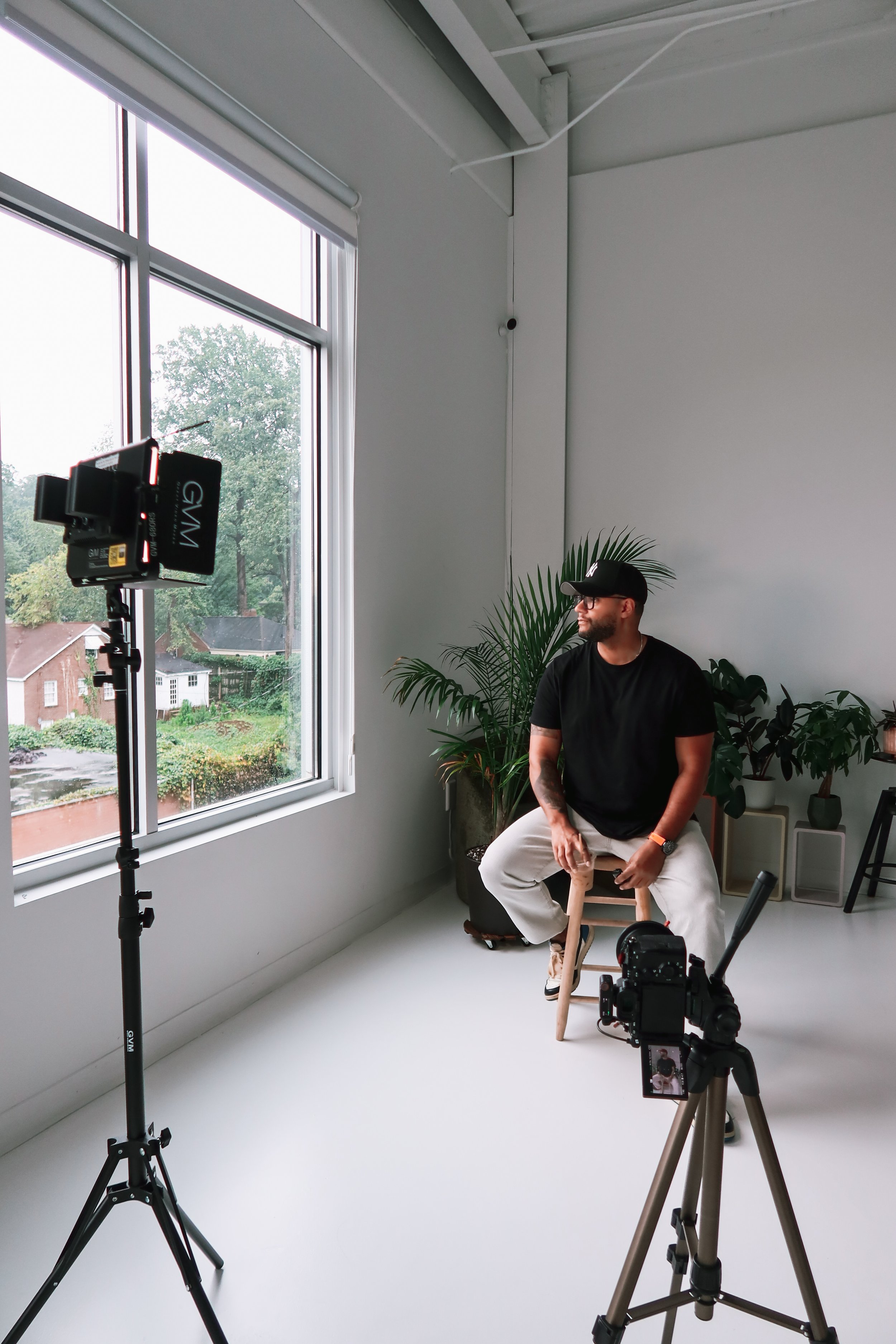 A man with a beard, wearing a black cap, black t-shirt, and beige pants, sitting on a stool in a photography studio by large window with trees and houses outside. He is surrounded by professional camera equipment, including a mounted camera and tripod, with a backdrop of green plants.