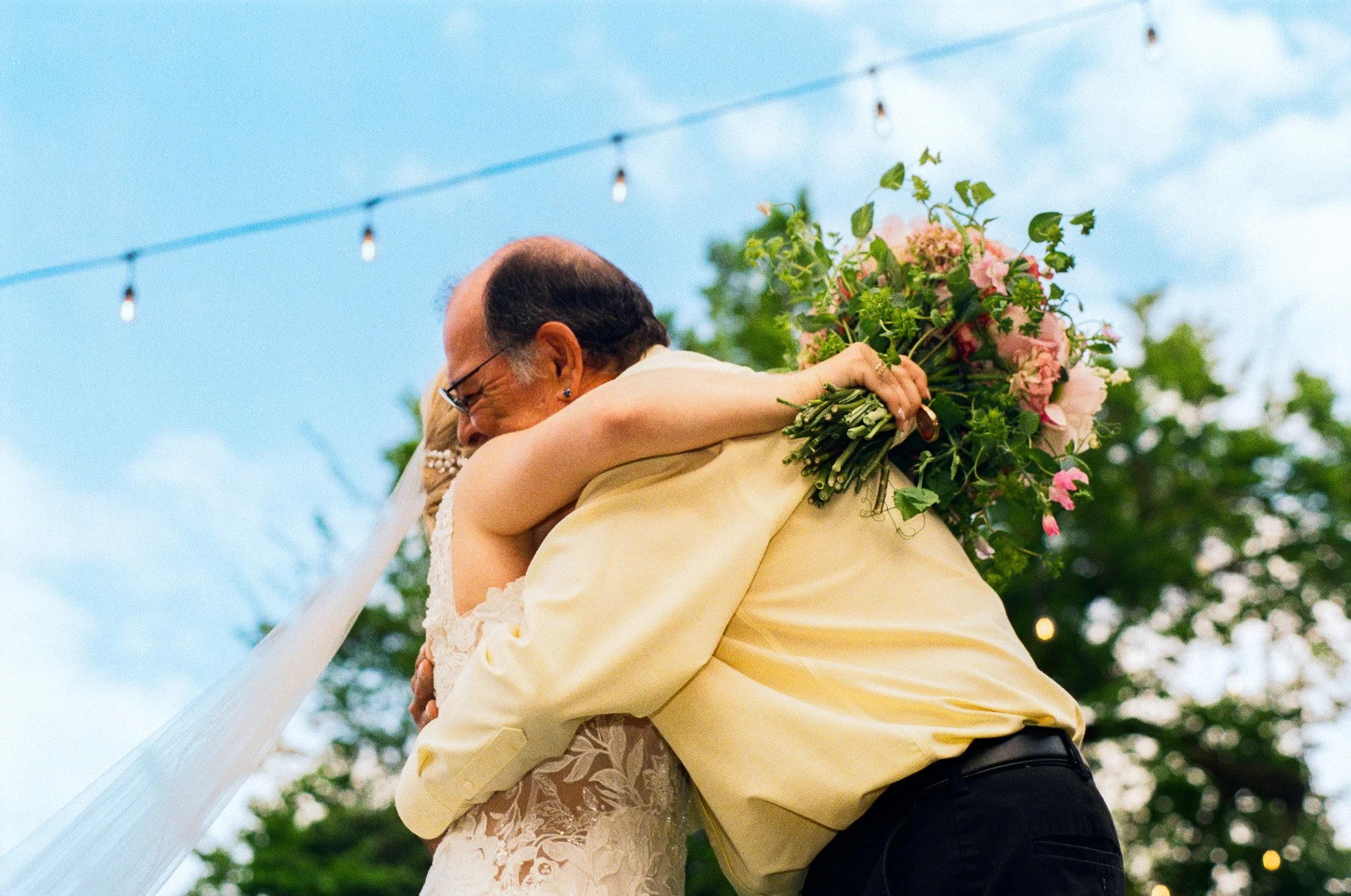 A couple hugging during a wedding ceremony outdoors, with a woman holding a bouquet of pink and green flowers, and string lights hanging above.