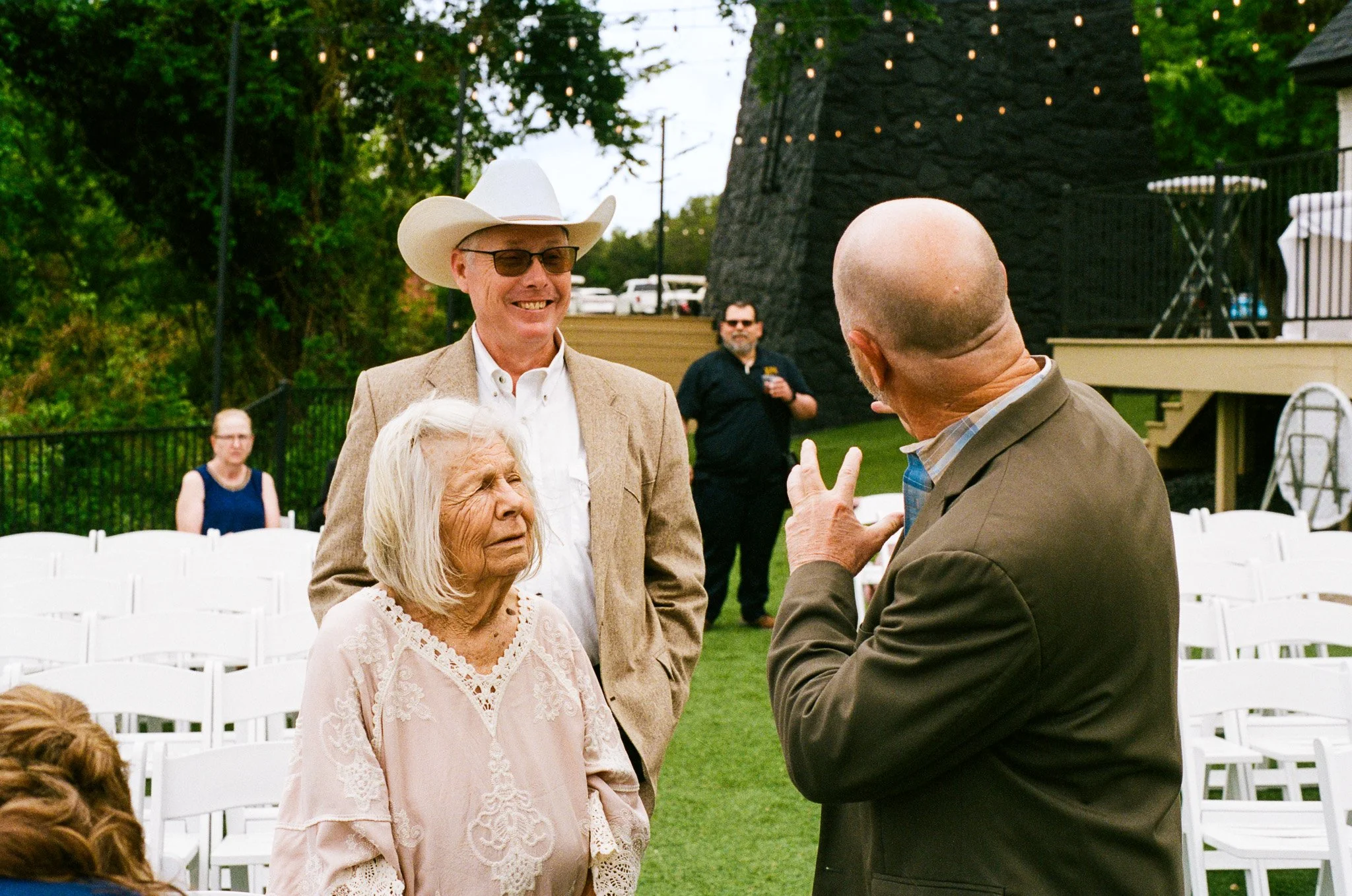 Group of elderly people and a man in a tan suit talking at an outdoor event with white chairs, string lights, and green trees in the background.