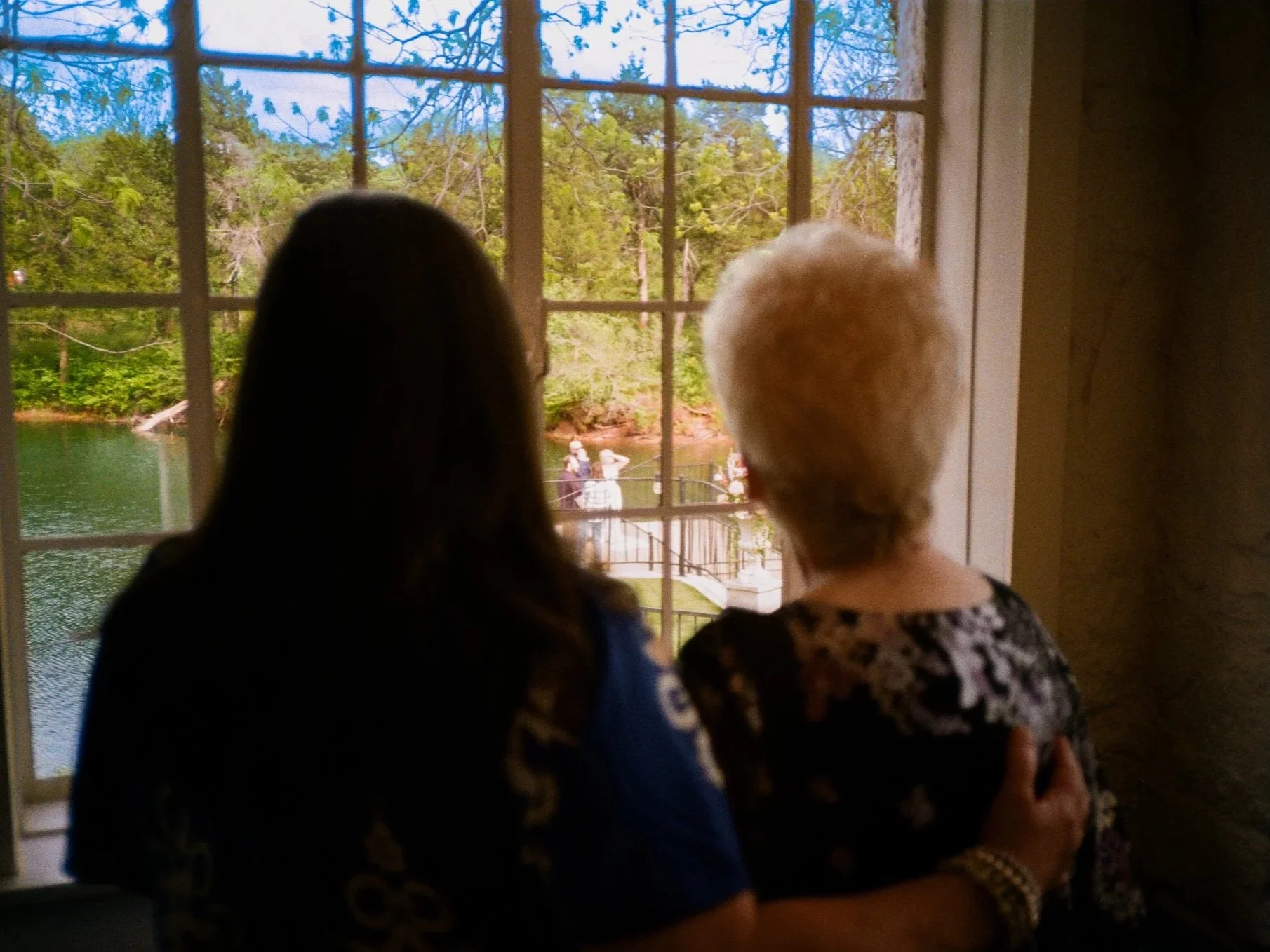 Two women, one with dark hair and the other with gray hair, are looking out a window at a peaceful outdoor scene with a wedding occurring outside.