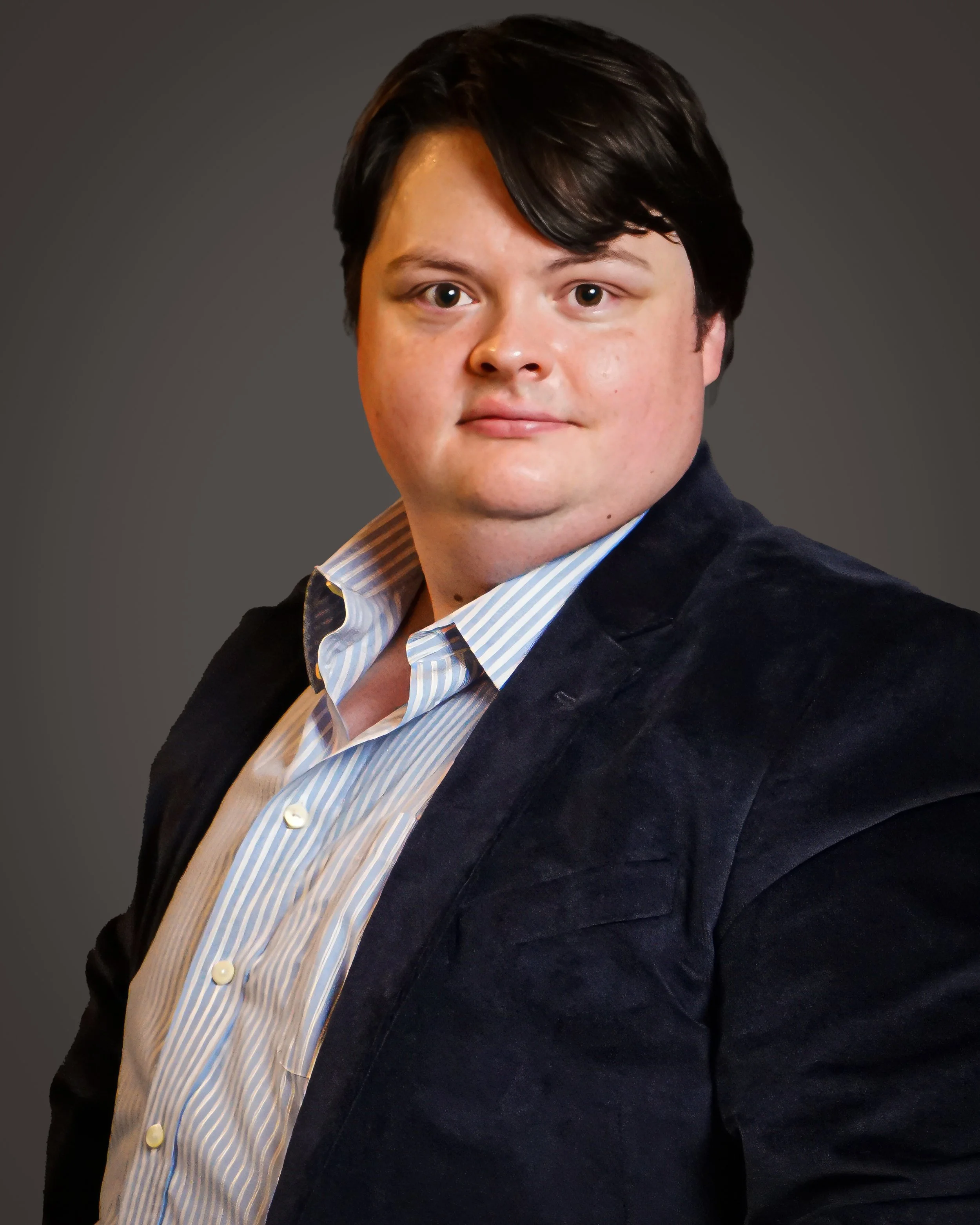 Portrait of a young man with black hair, wearing a striped shirt and a dark blazer, looking at the camera with a neutral expression against a gray background.