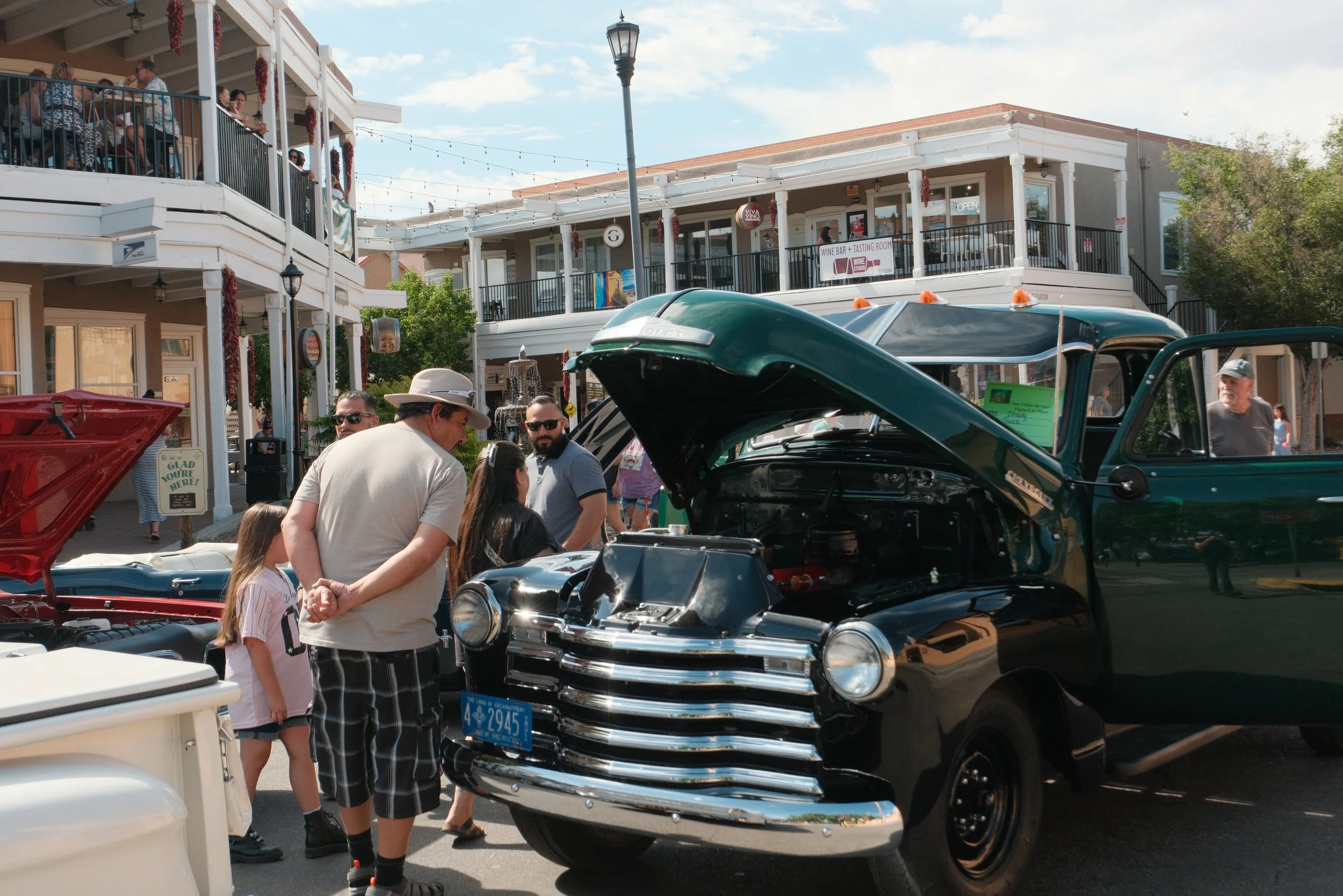 A street scene with vintage cars on display and people gathered around, some looking under the hoods of the cars. Two-story buildings with shops and restaurants, including a wine bar and tasting room, are visible in the background, along with a clear