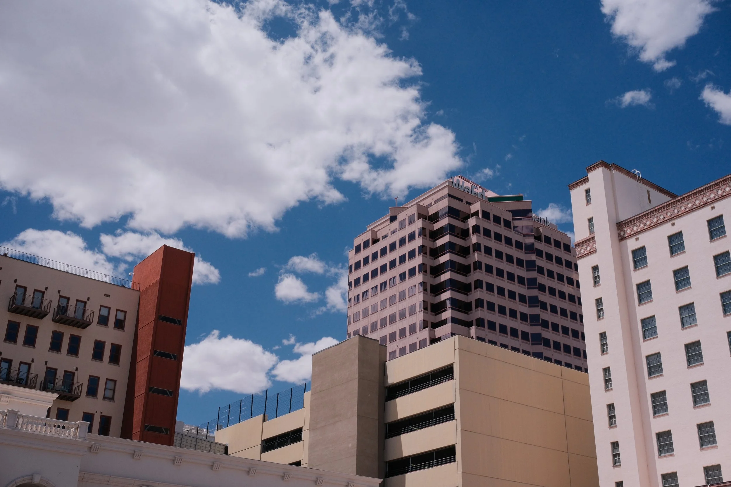 Various tall buildings in an urban area with a bright blue sky and scattered white clouds above.