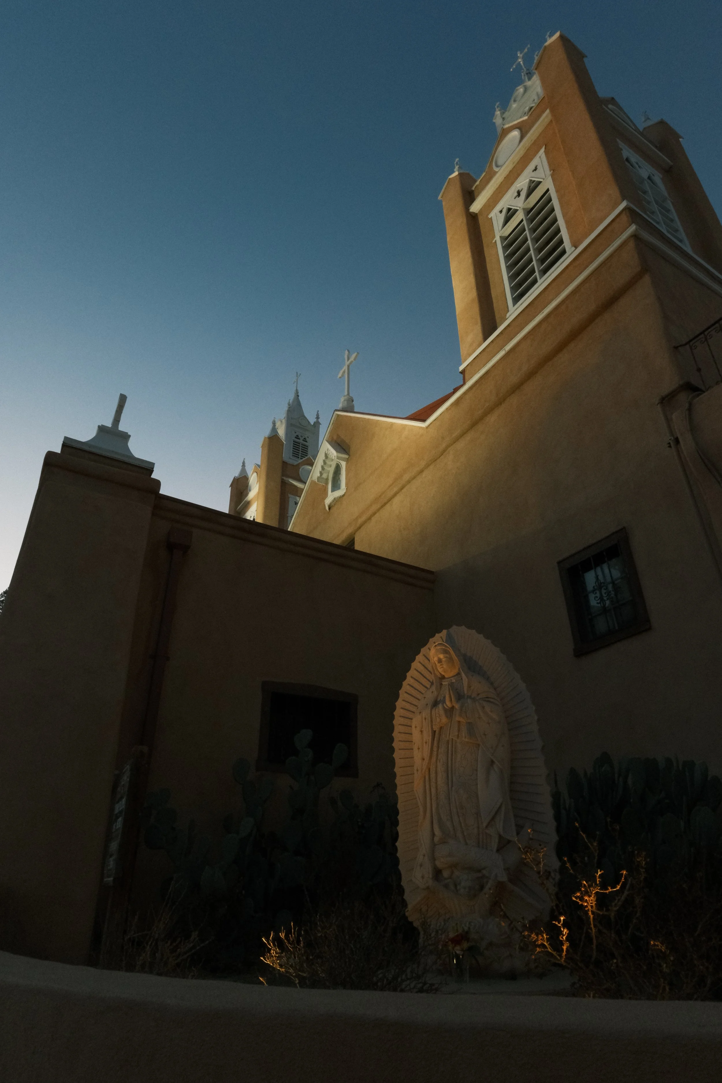 A church with towers and crosses, and a religious statue at the entrance, illuminated at night with a clear dark blue sky in the background.